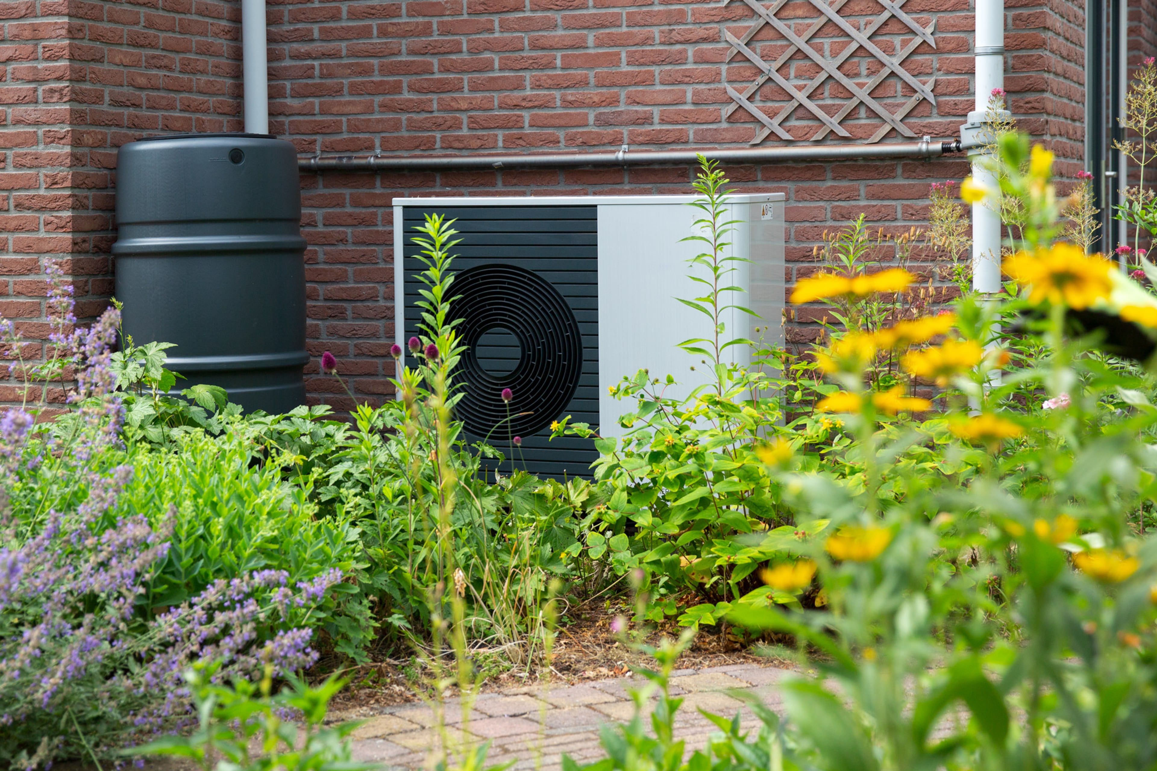 Buitenunit van een warmtepomp. Ernaast staat een regenton, ervoor gele bloemen
