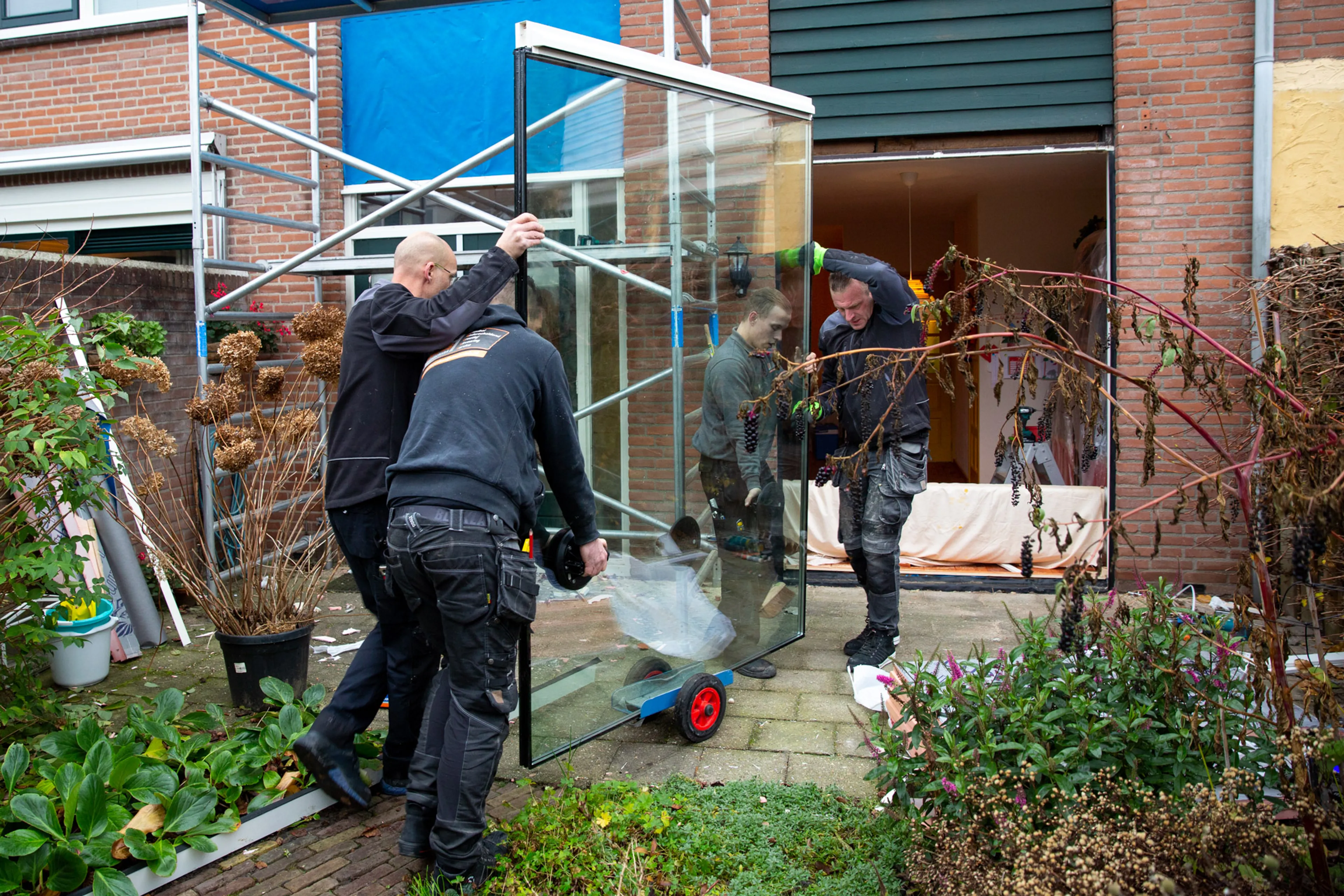 Vier mannen rijden grote glasplaat met triple glas naar het raam van de woonkamer.