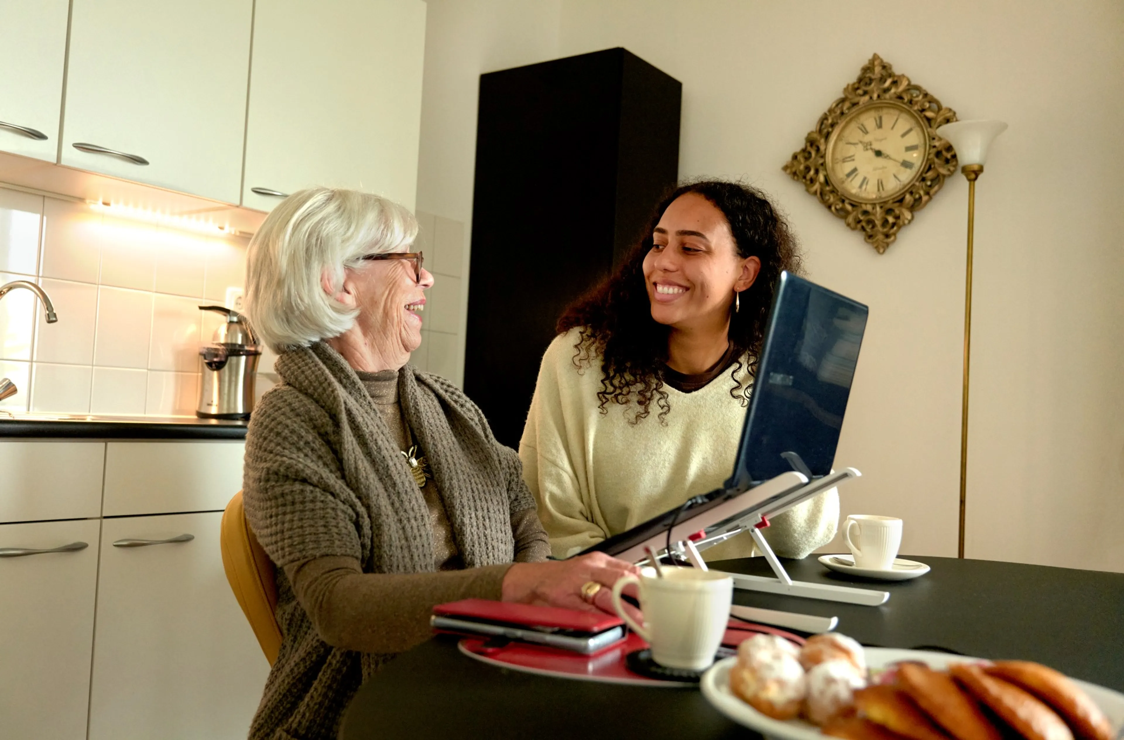 Twee vrouwen zitten in de keuken achter een laptop. Ze lachen naar elkaar.