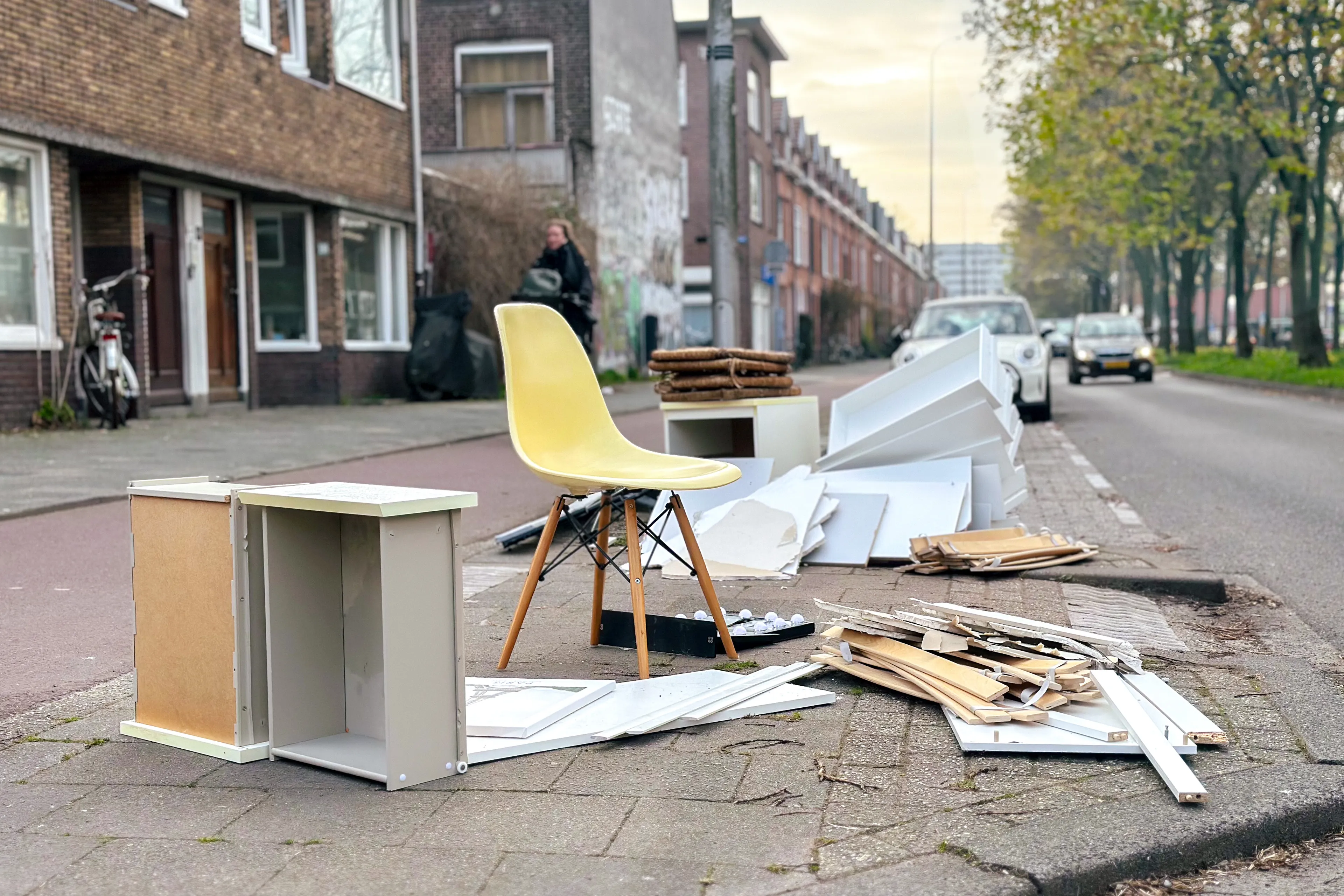 Op de afbeelding zie je een kast die uit elkaar gehaald is, een stoel en een lattenbodem op de stoep naast een weg.