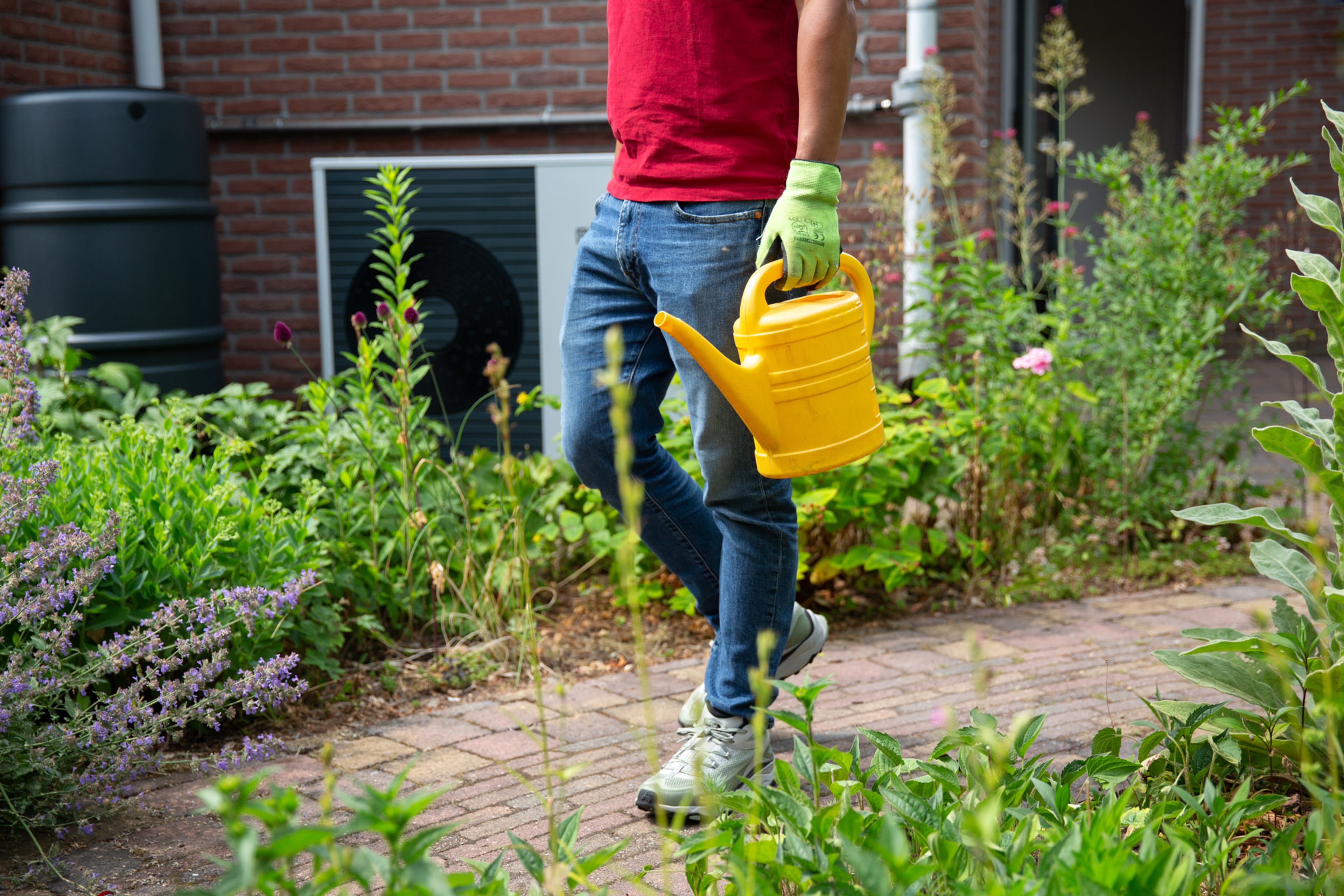 Buitenunit van een warmtepomp. Ervoor loopt een man met gele gieter