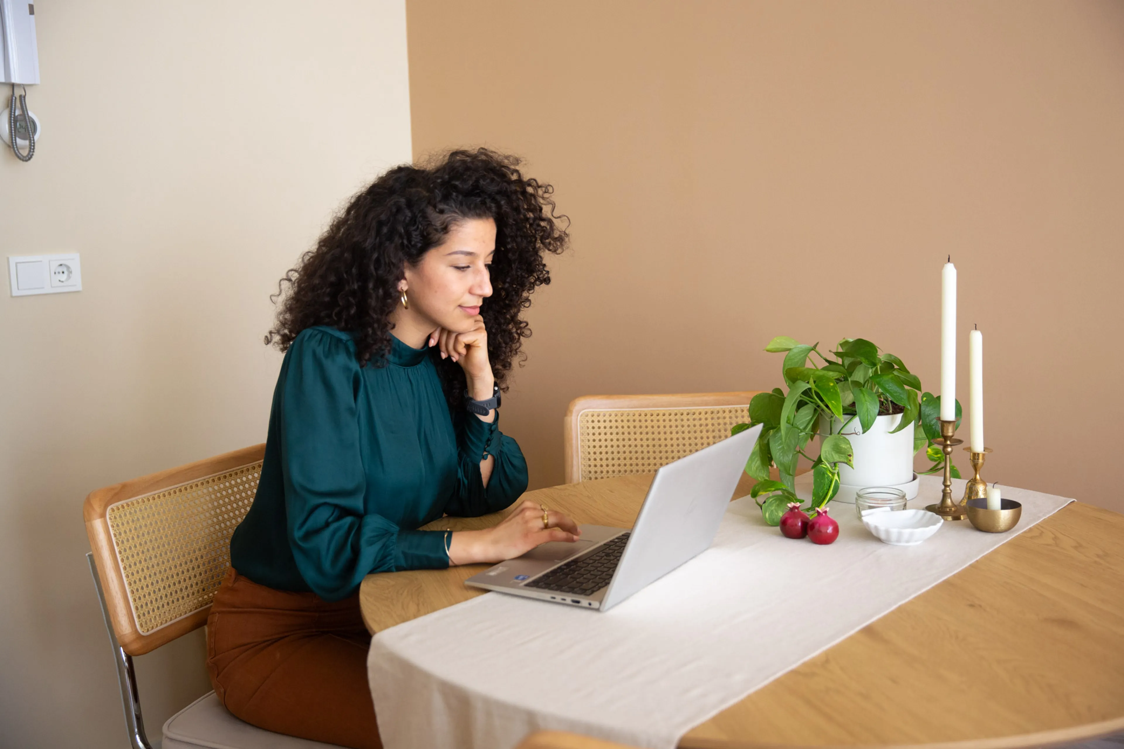Een vrouw met zwart krulhaar en een donkergroene blouse zit aan een tafel met een laptop.