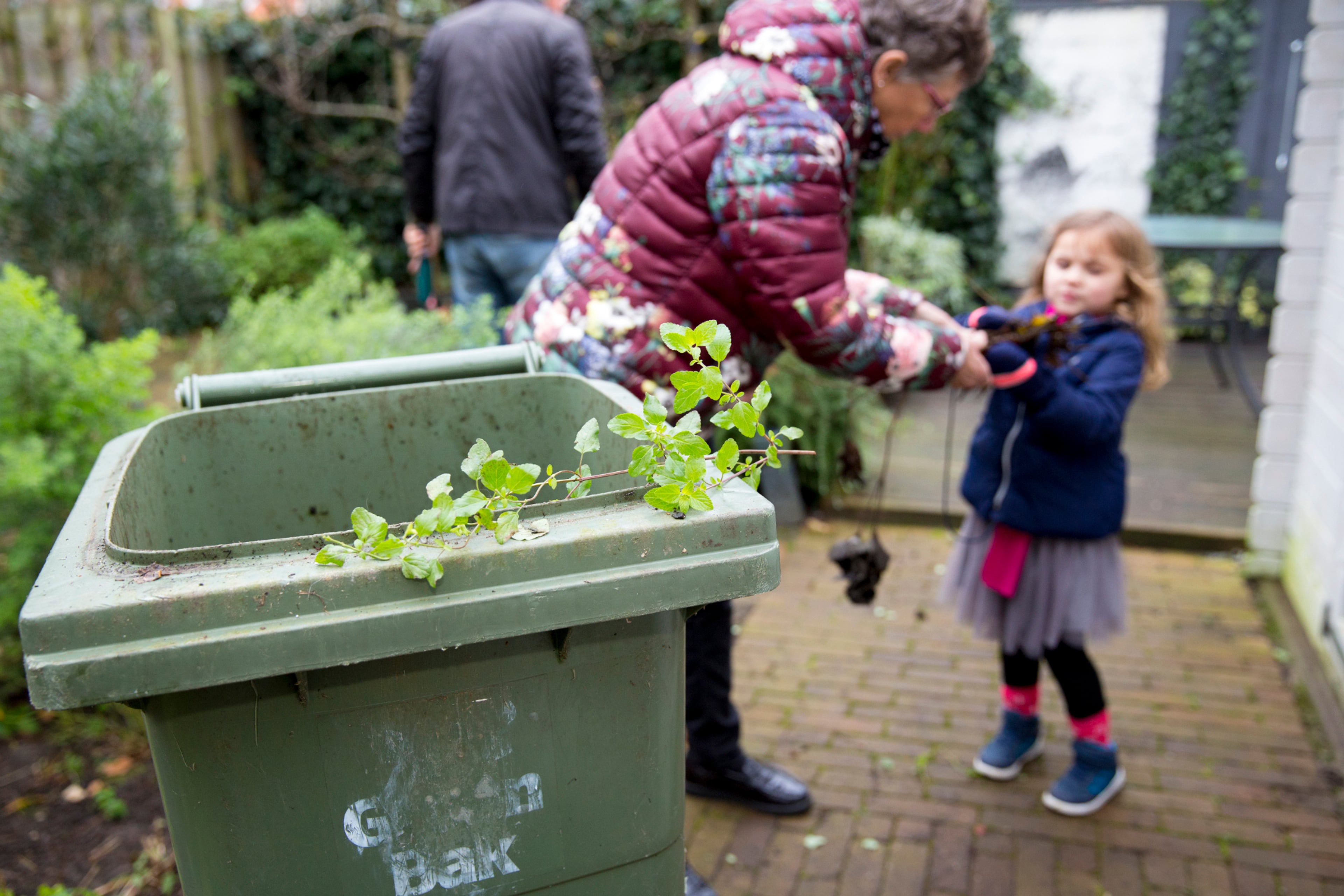 Vrouw en meisje in de tuin met een GFT bak op de voorgrond. Vraag een GFT bak aan bij de gemeente, wanneer je deze nog niet hebt.
