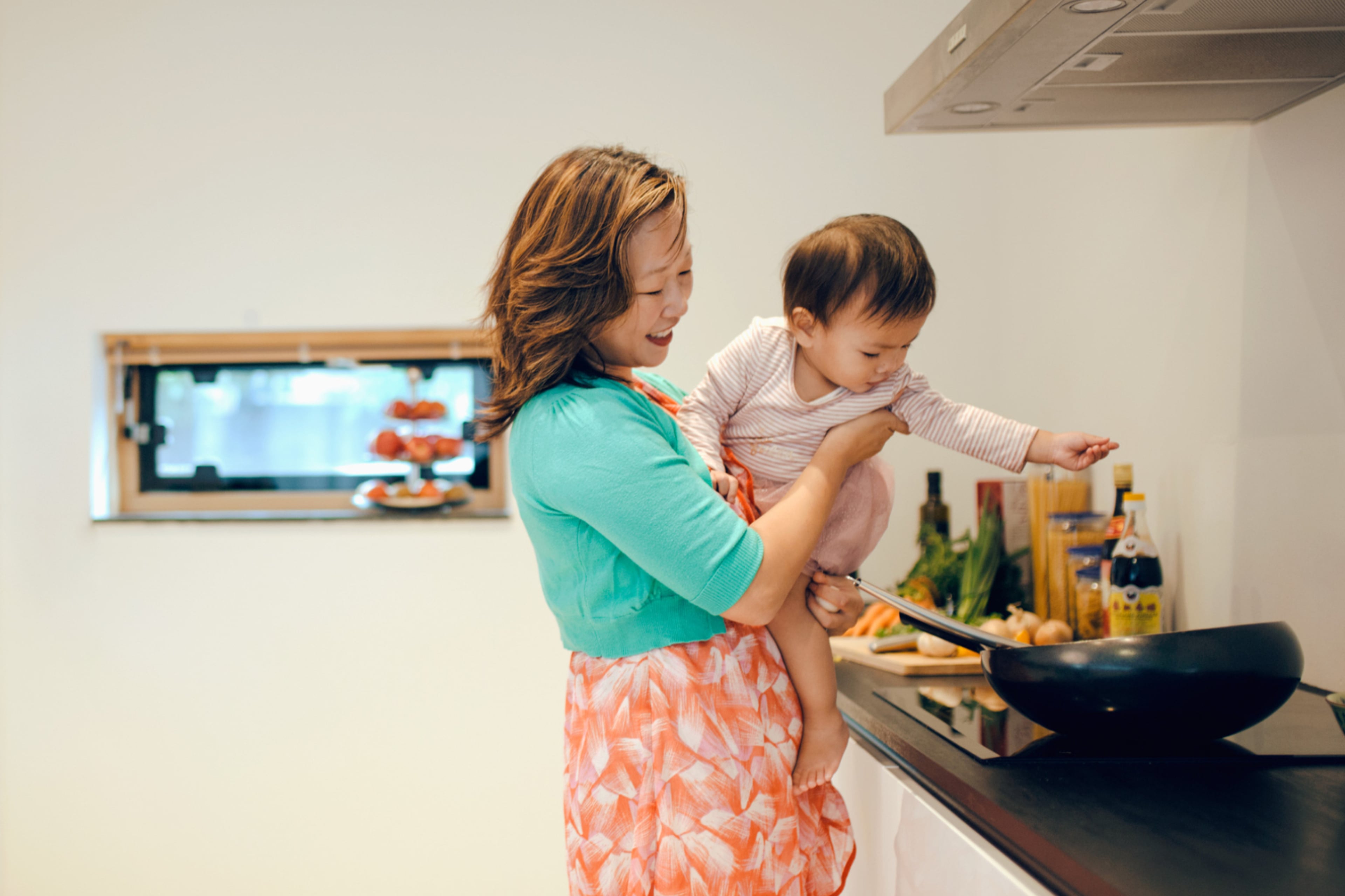 Vrouw staat bij fornuis met kind in de armen. Kies voor een inductie kookplaat wanneer je een nieuwe keuken aanschaft.