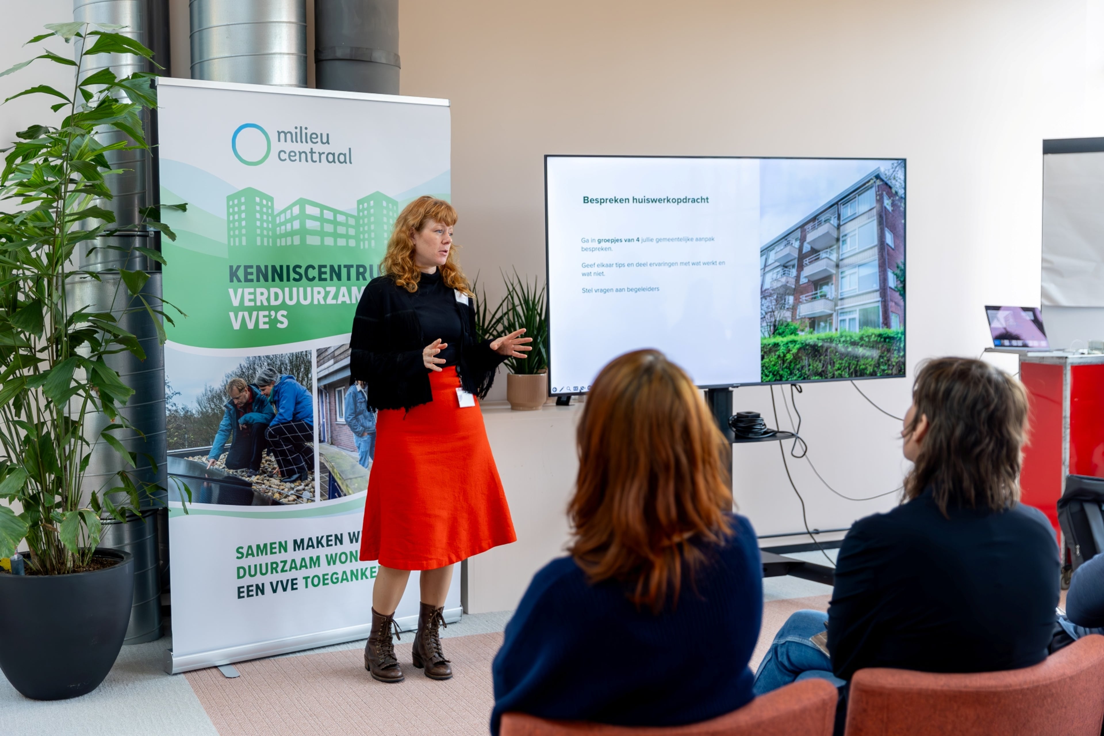 Een vrouw met rossig haar en een rode rok spreekt een groep mensen toe, voor een banner waarop staat "Kenniscentrum Verduurzaming VvE's"