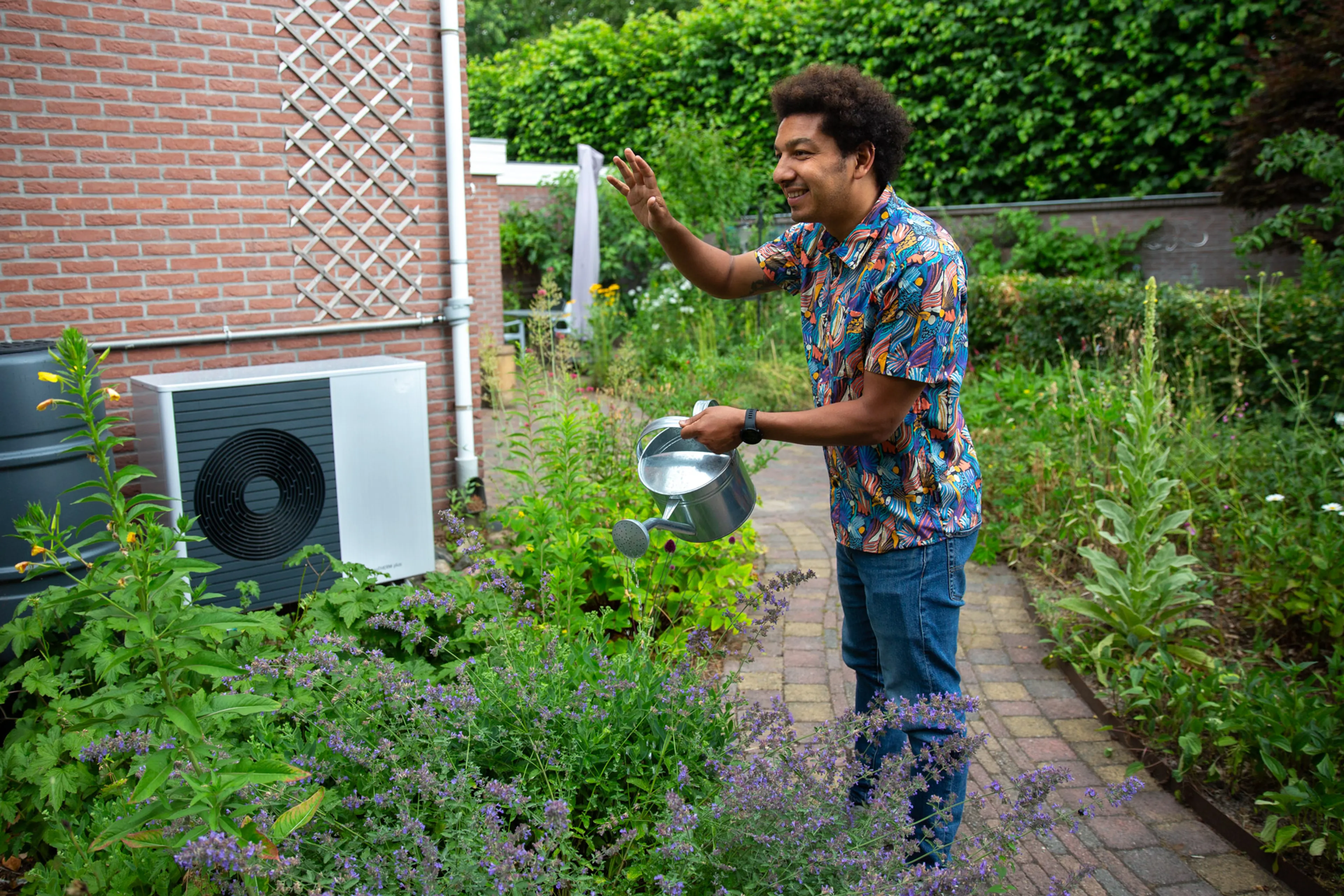 Buitenunit van een warmtepomp. Ervoor staat een man met een gieter.