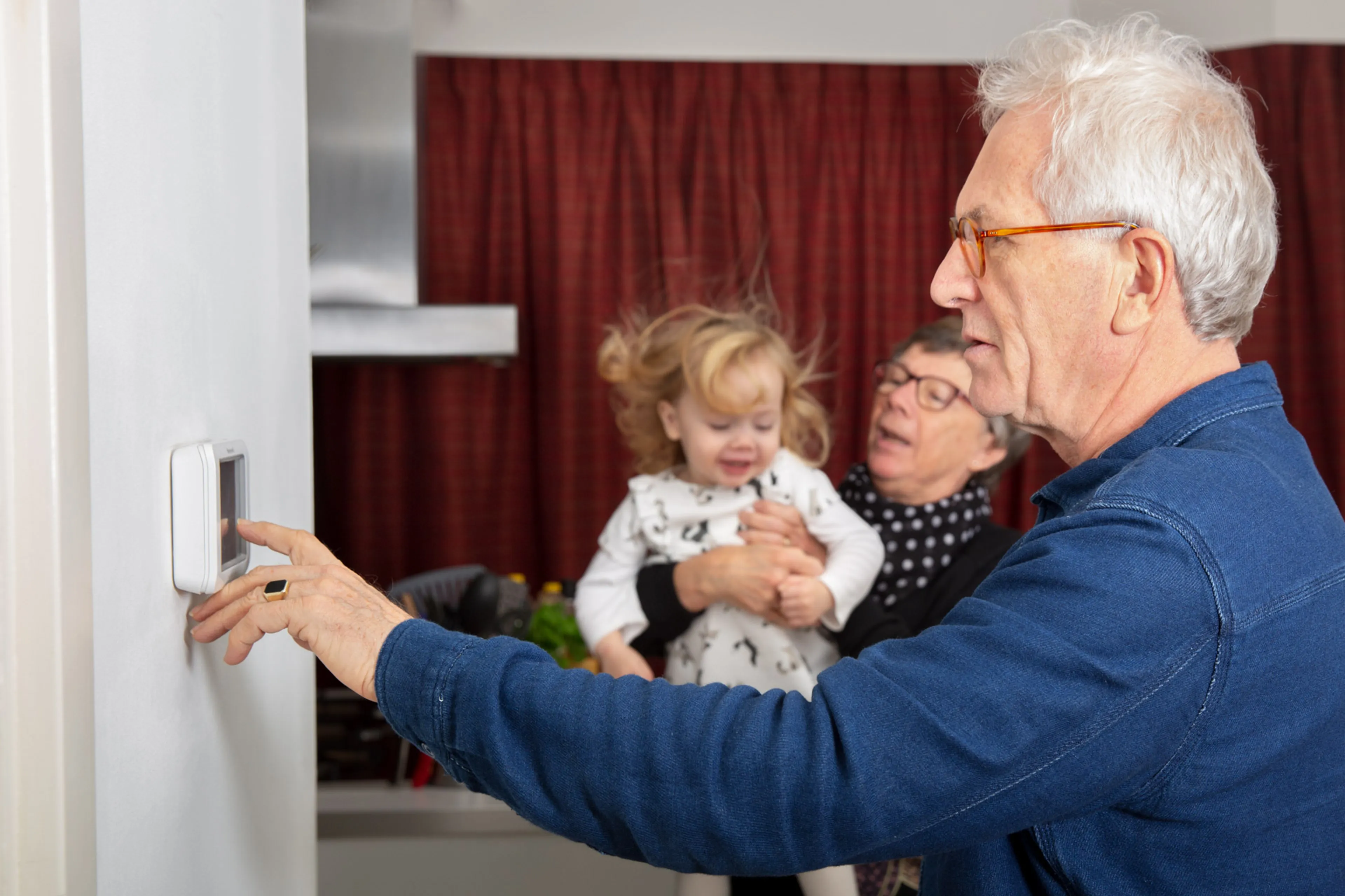 Opa bij kamerthermostaat, oma met kleindochter op de achtergrond. Bespaar energie door de thermostaat een graad lager te zetten.