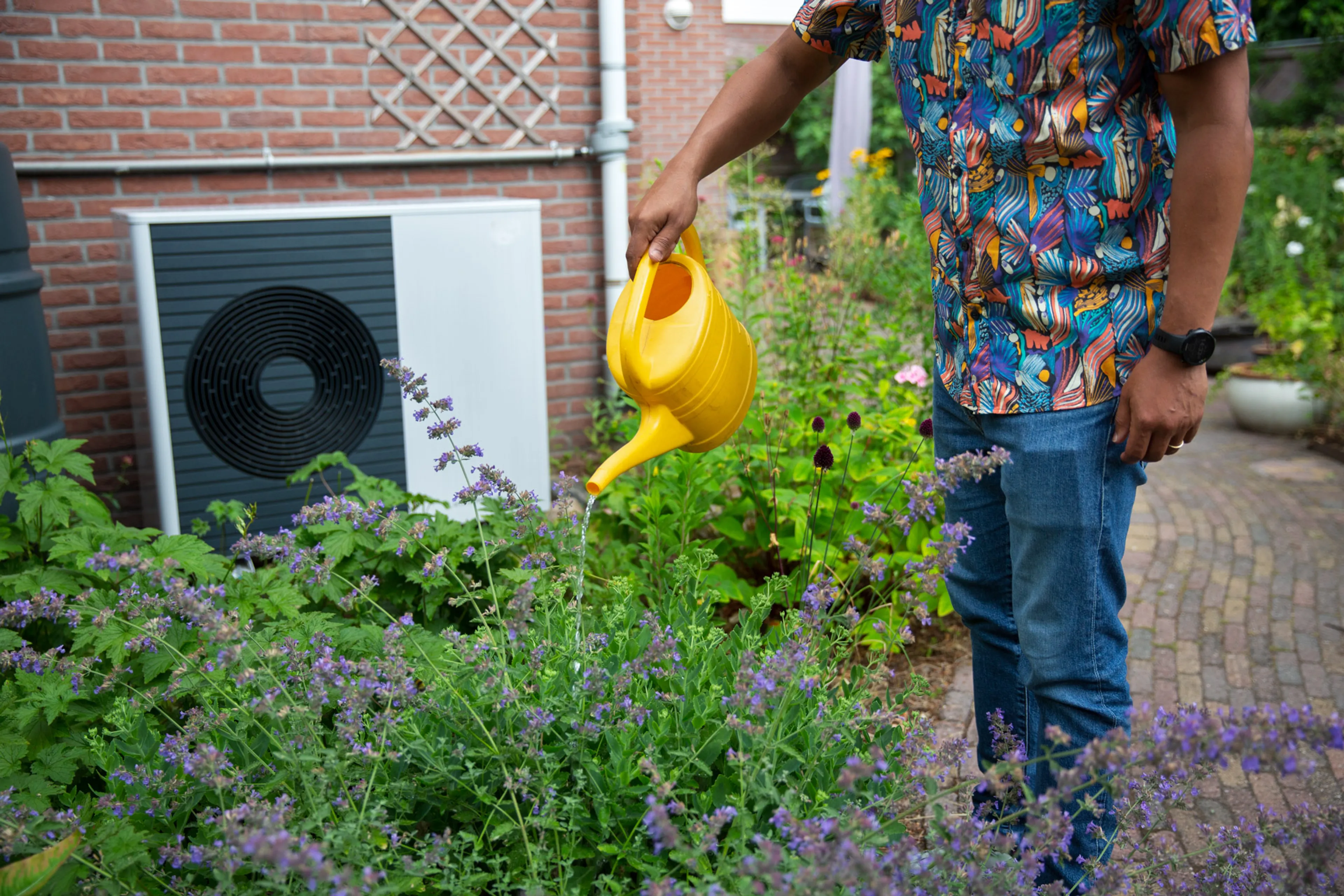 Buitenunit van een warmtepomp. Ervoor staat een man met een gele gieter.