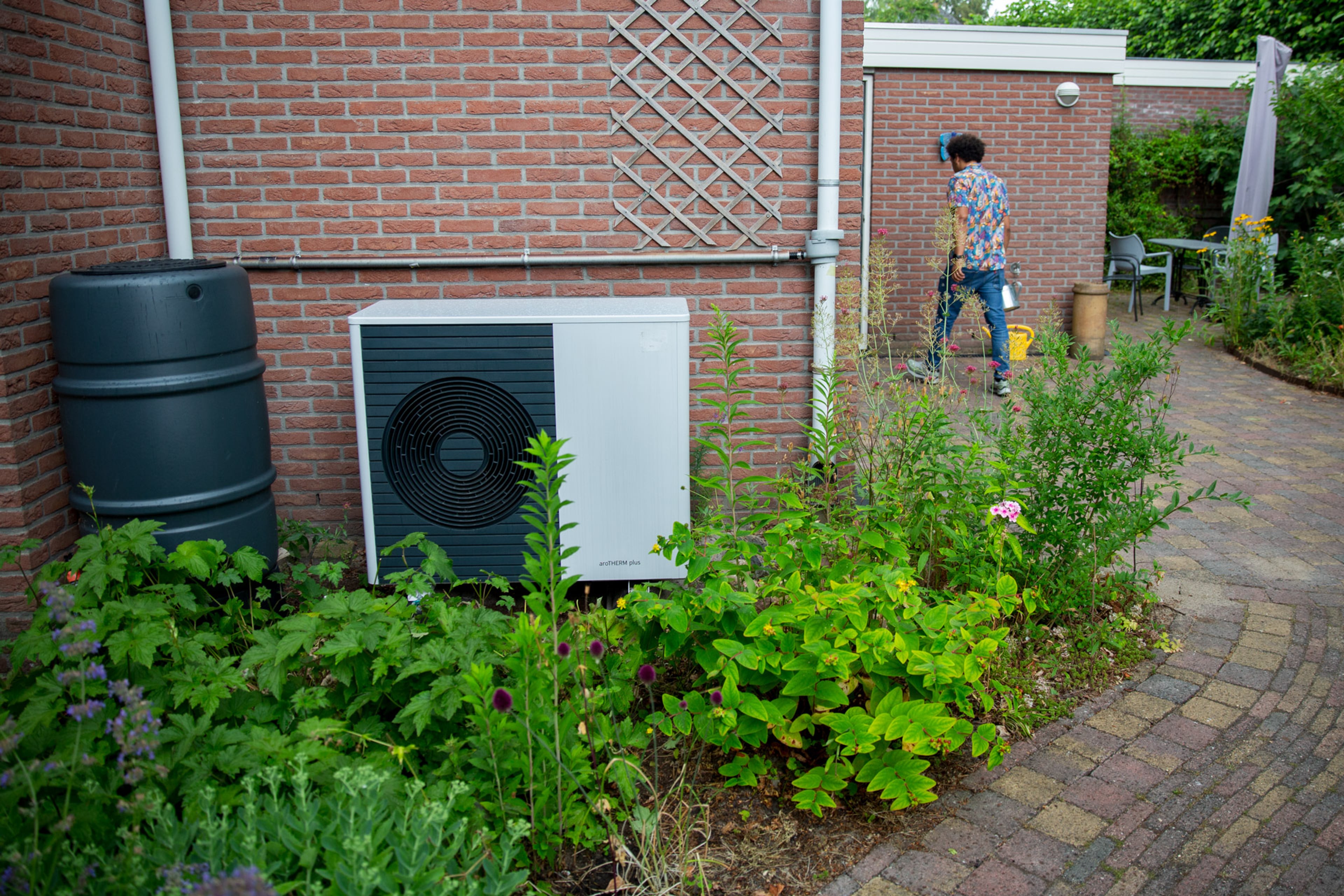 Buitenunit van een warmtepomp. Op de achtergrond loopt een man met een gieter.