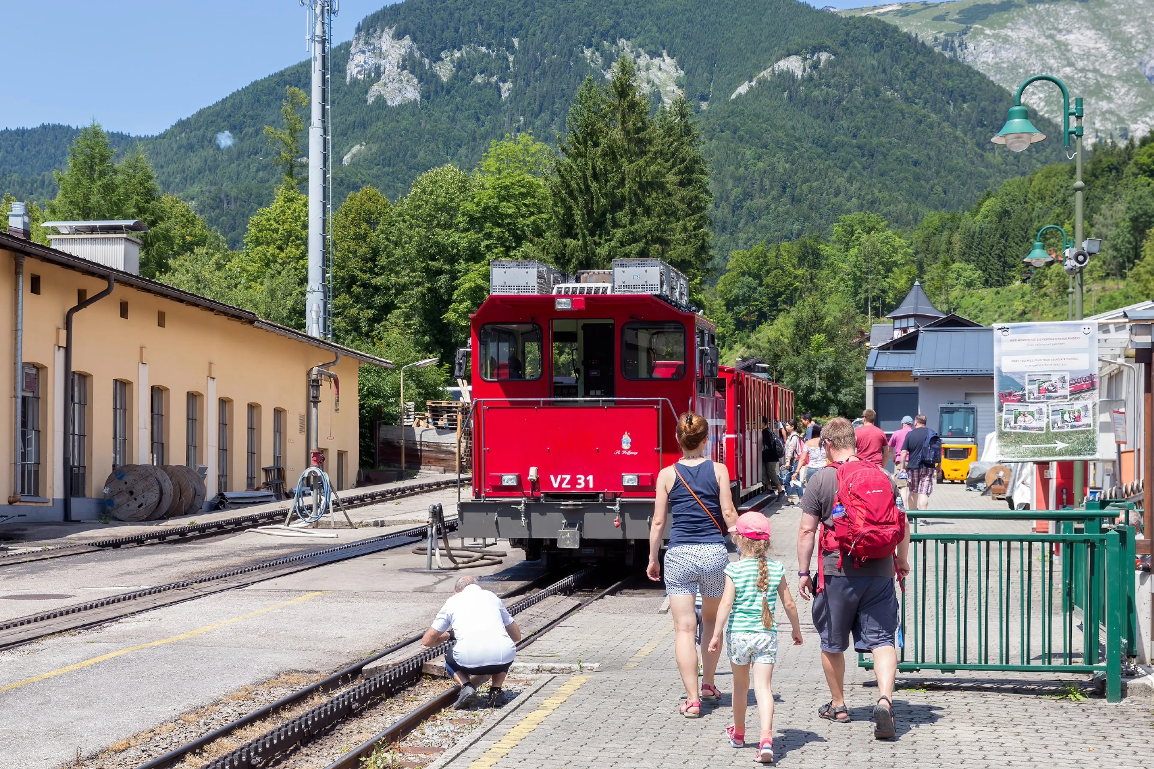 Een gezin loopt richting een rode trein in de Oostenrijkse bergen. Een gezinsvakantie met de trein is vaak makkelijk en goedkoop.