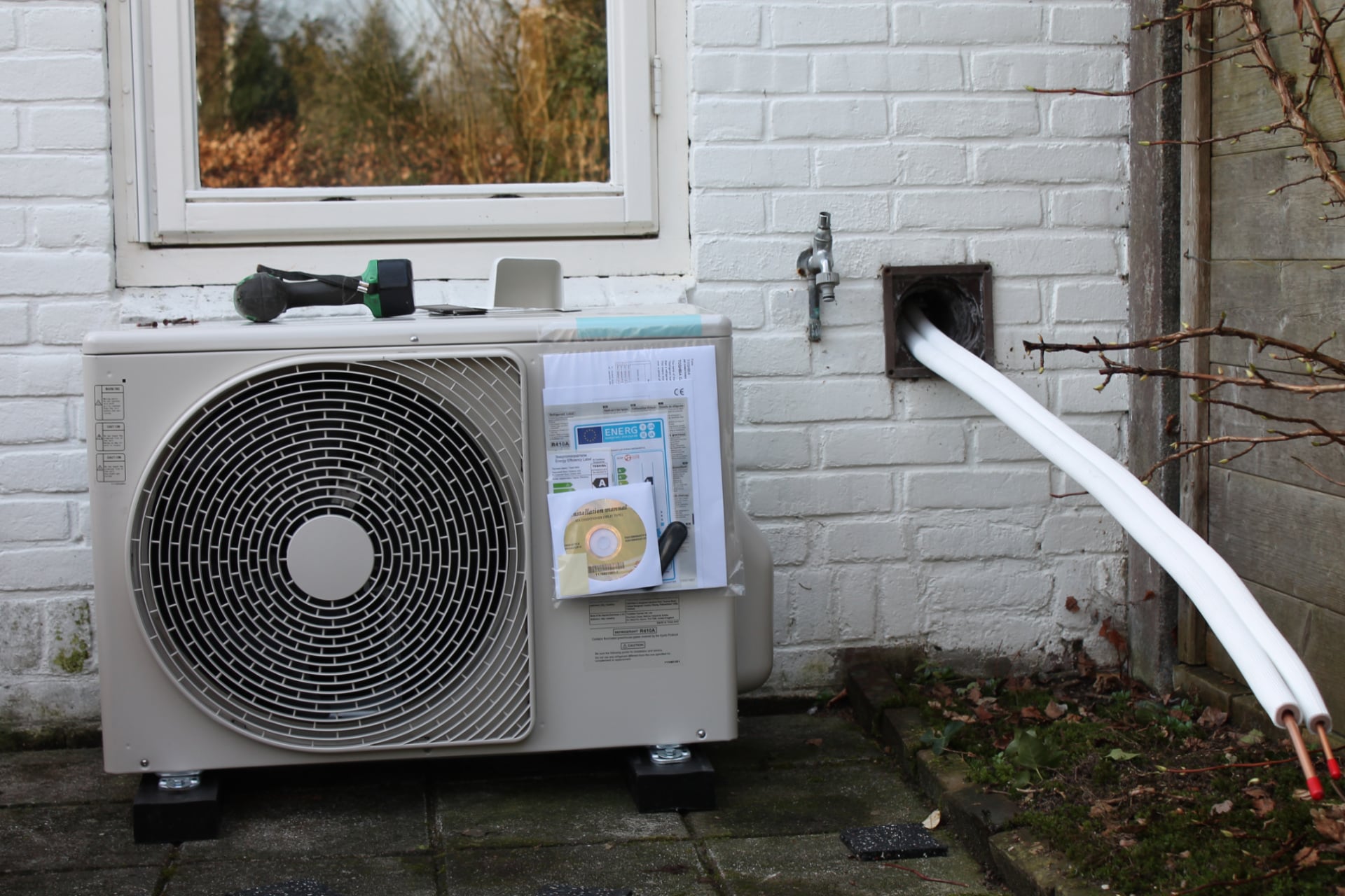 Buitenunit van hybride warmtepomp tegen buitenmuur onder raam.