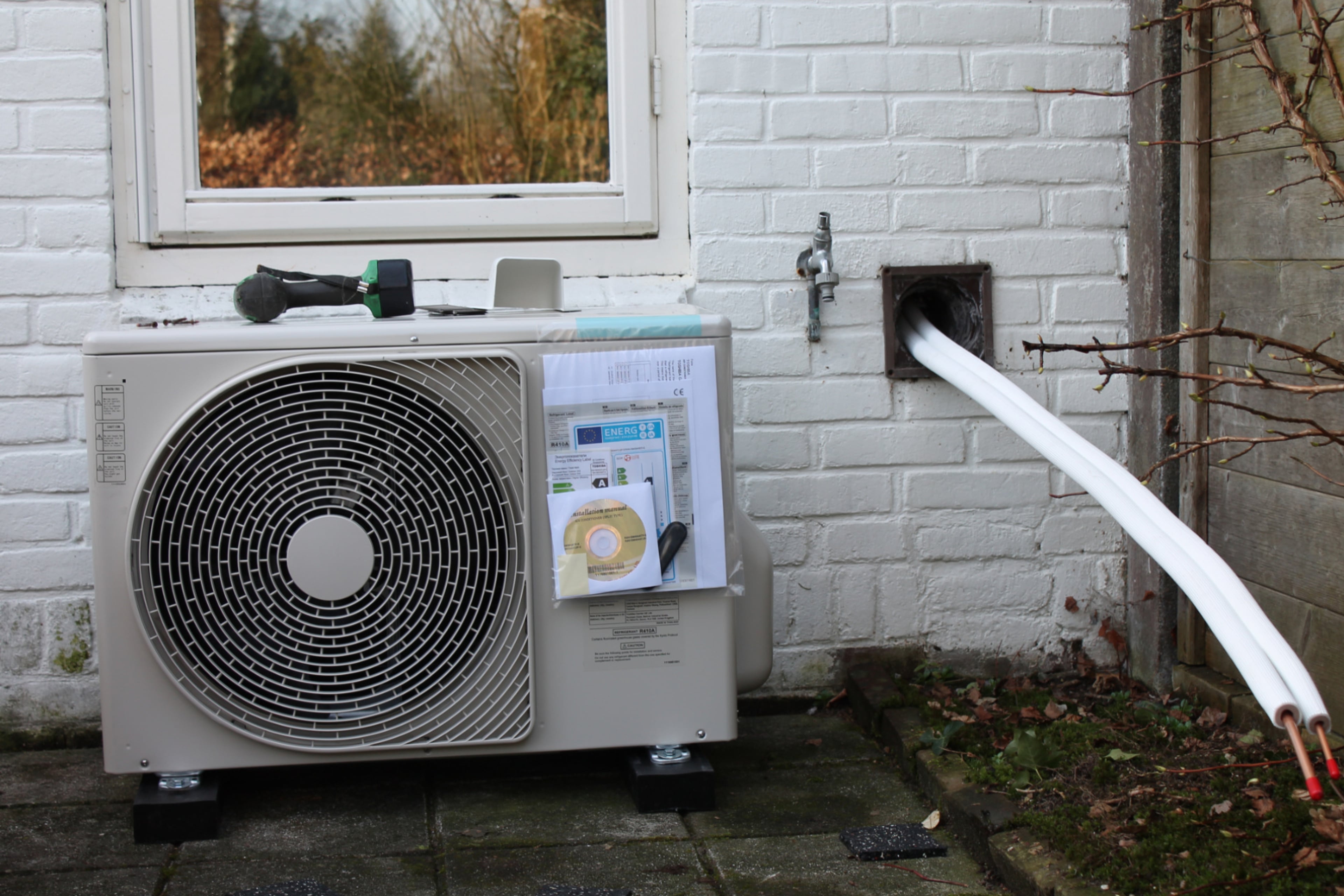 Buitenunit van hybride warmtepomp tegen buitenmuur onder raam.