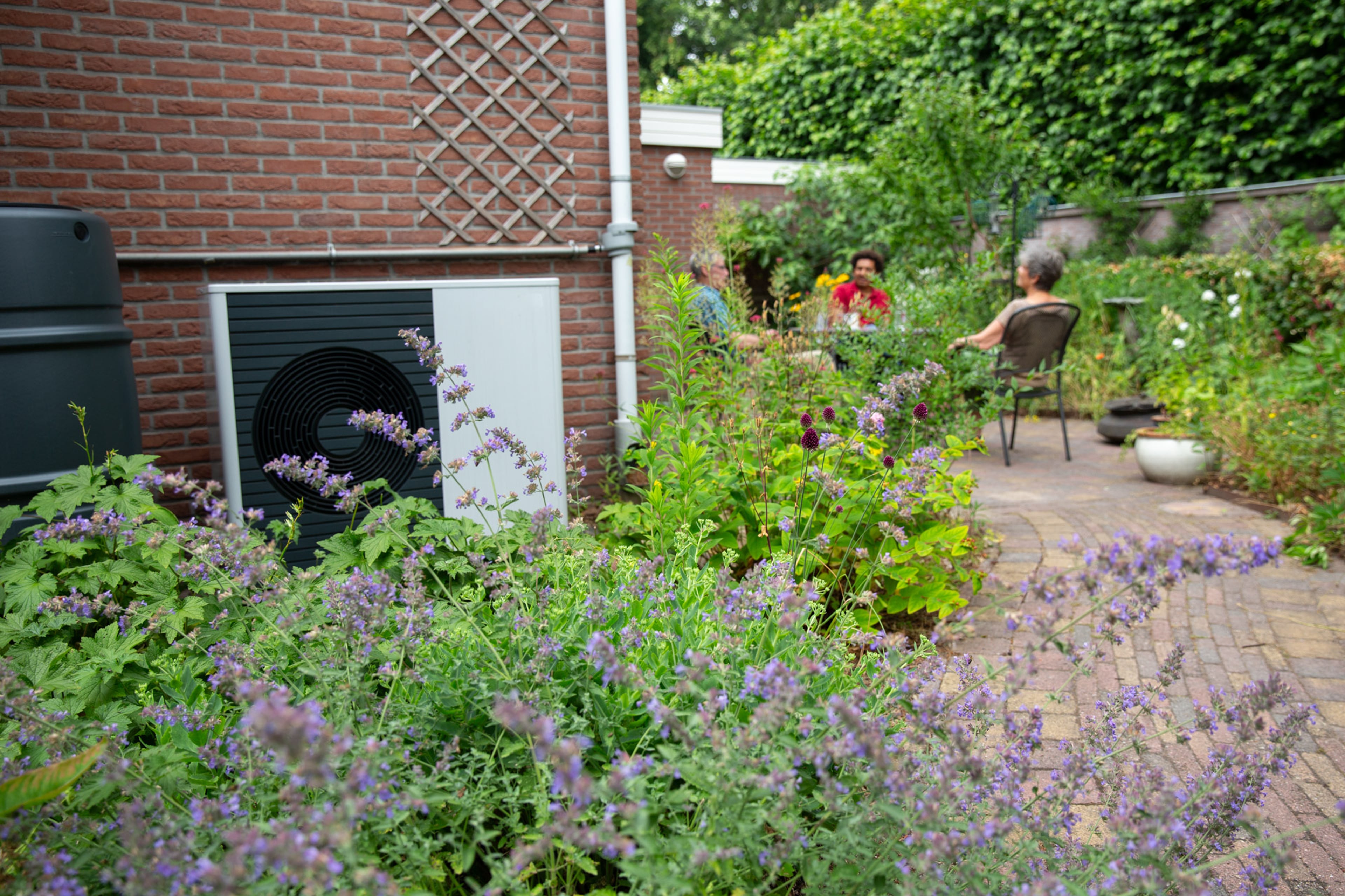 Buitenunit van een warmtepomp tegen de muur van het huis. Op het terras erachter zitten mensen.