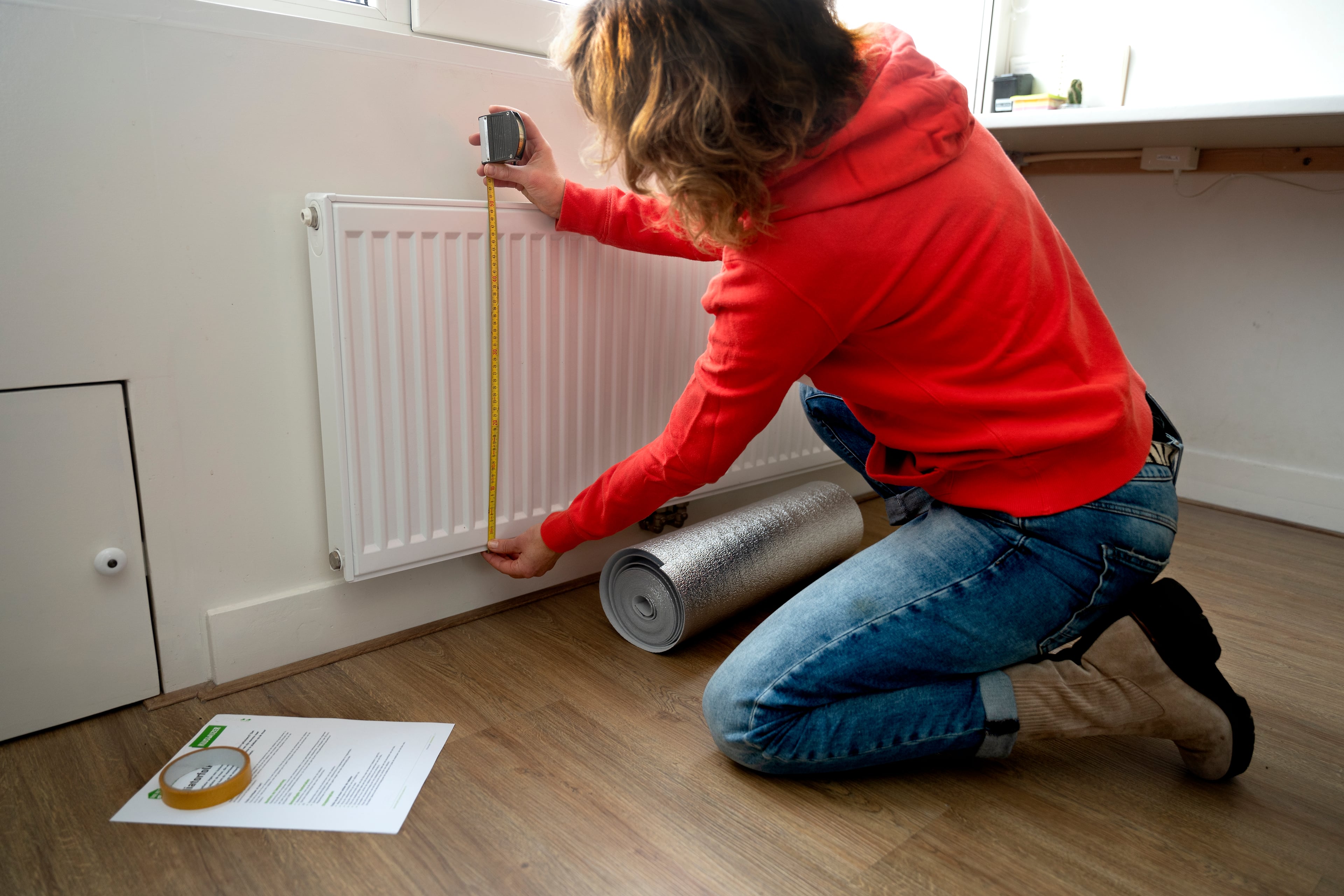 Vrouw met meetlint bij radiator. Ze meet de radiator op zodat ze de radiatorfolie op maat kan knippen