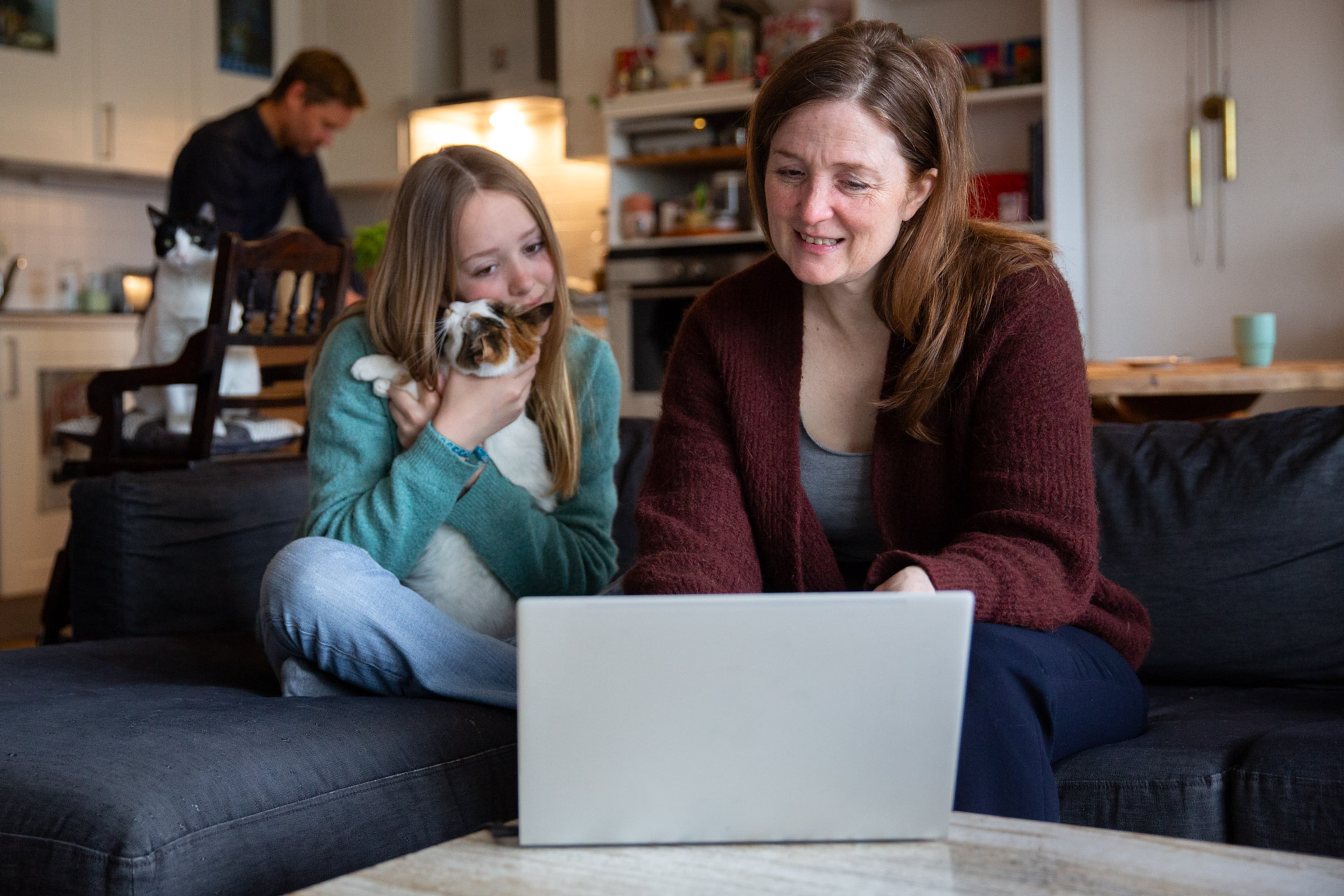 Een vrouw zit met haar dochter op de bank. Ze kijken naar een laptop, het meisje heeft een kat op schoot.