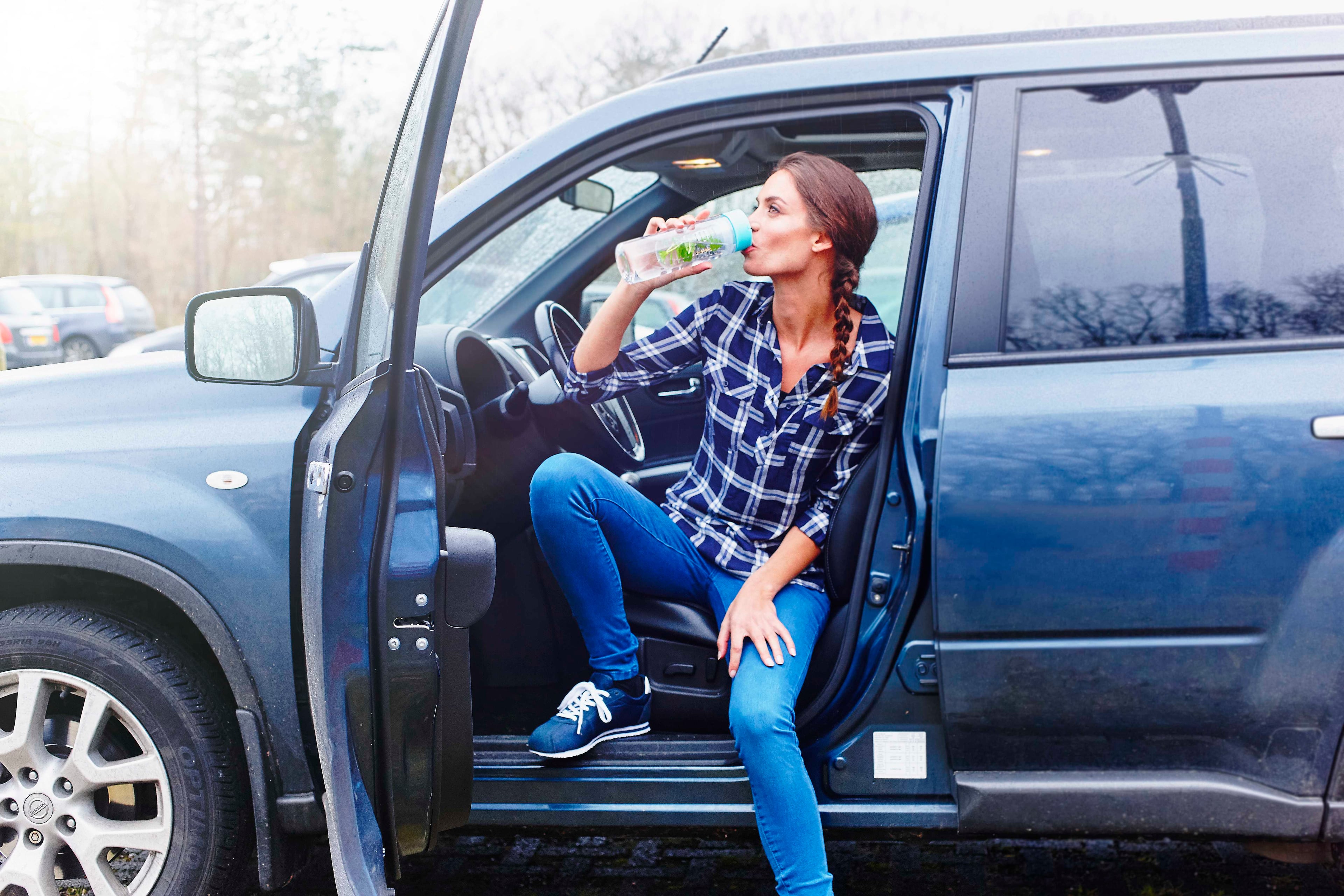 Vrouw drinkt zitten in portier van de auto uit een meeneemfles. Voorkom (plastic) afval en recycle beter.