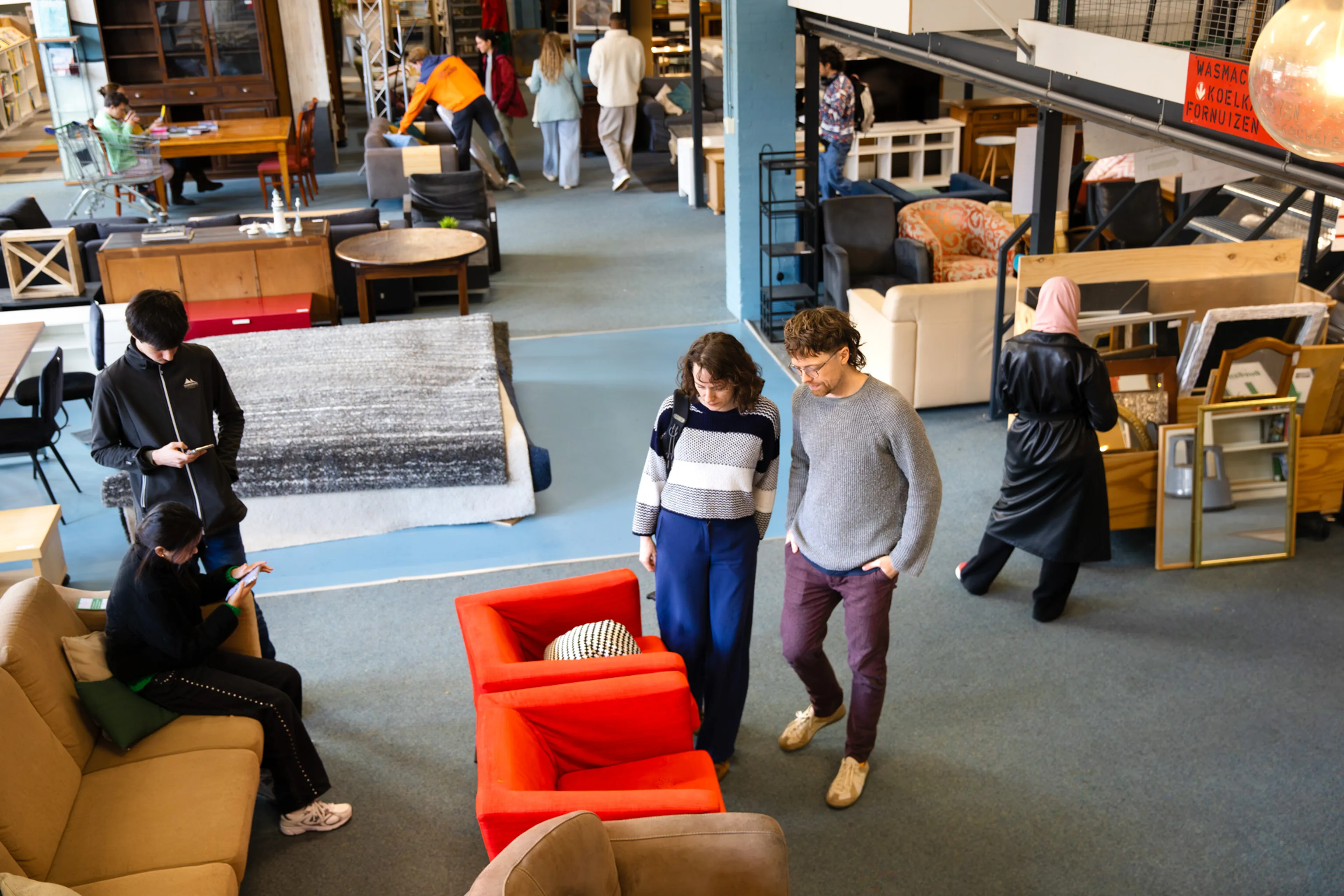 Op de afbeelding zie je een kringloopwinkel. Er staan verschillende meubels, zoals kasten, banken en stoelen. In de winkel lopen mensen rond die de meubels bekijken.