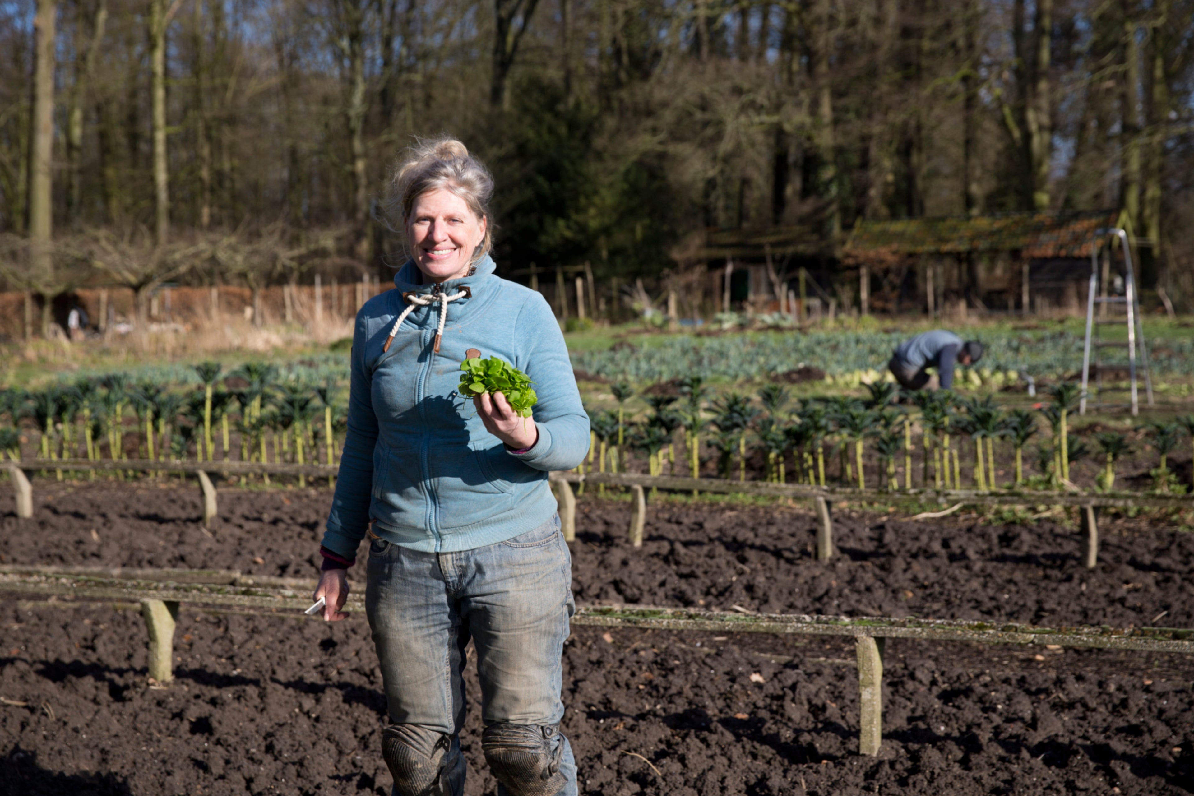 Vrouw poseert lachend in een biologische moestuin met een gewas in haar handen. Biologische landbouw versus gangbare landbouw.