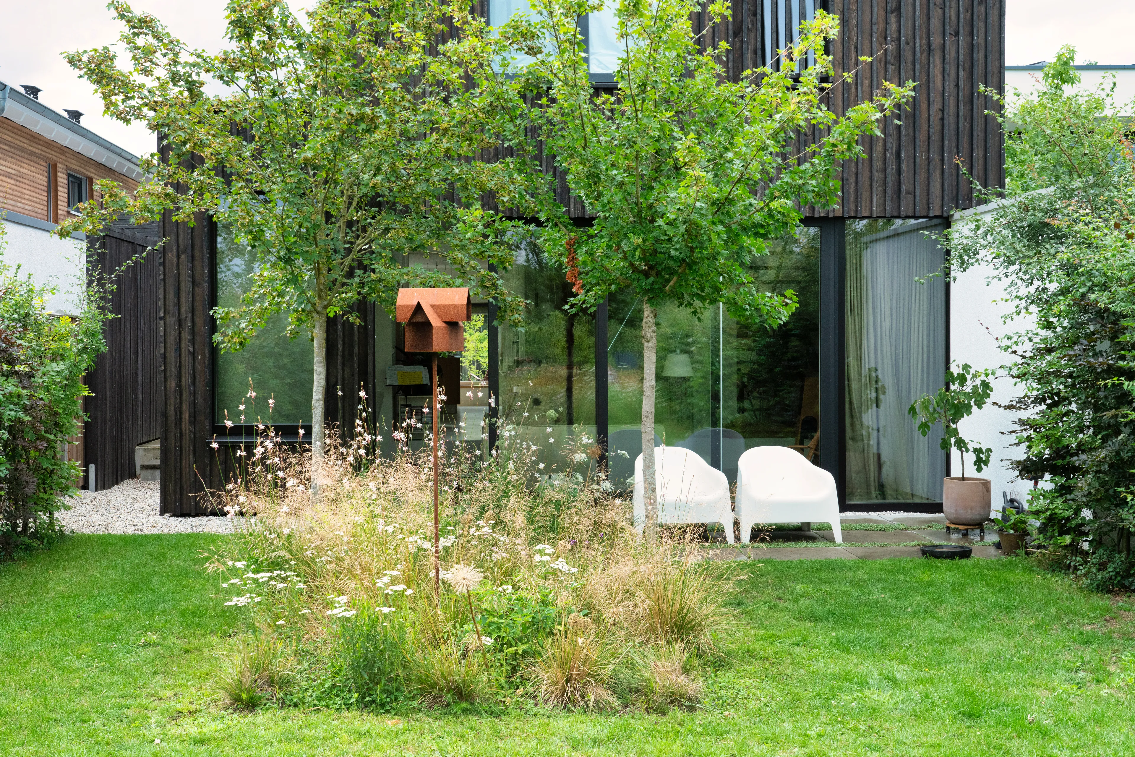 Op de afbeelding zie je het gazon van een tuin. In het midden van het gazon is er een plekje met bloemen en planten en twee bomen. Op de achtergrond zie je de achtergevel van een huis en twee witte tuinstoelen.