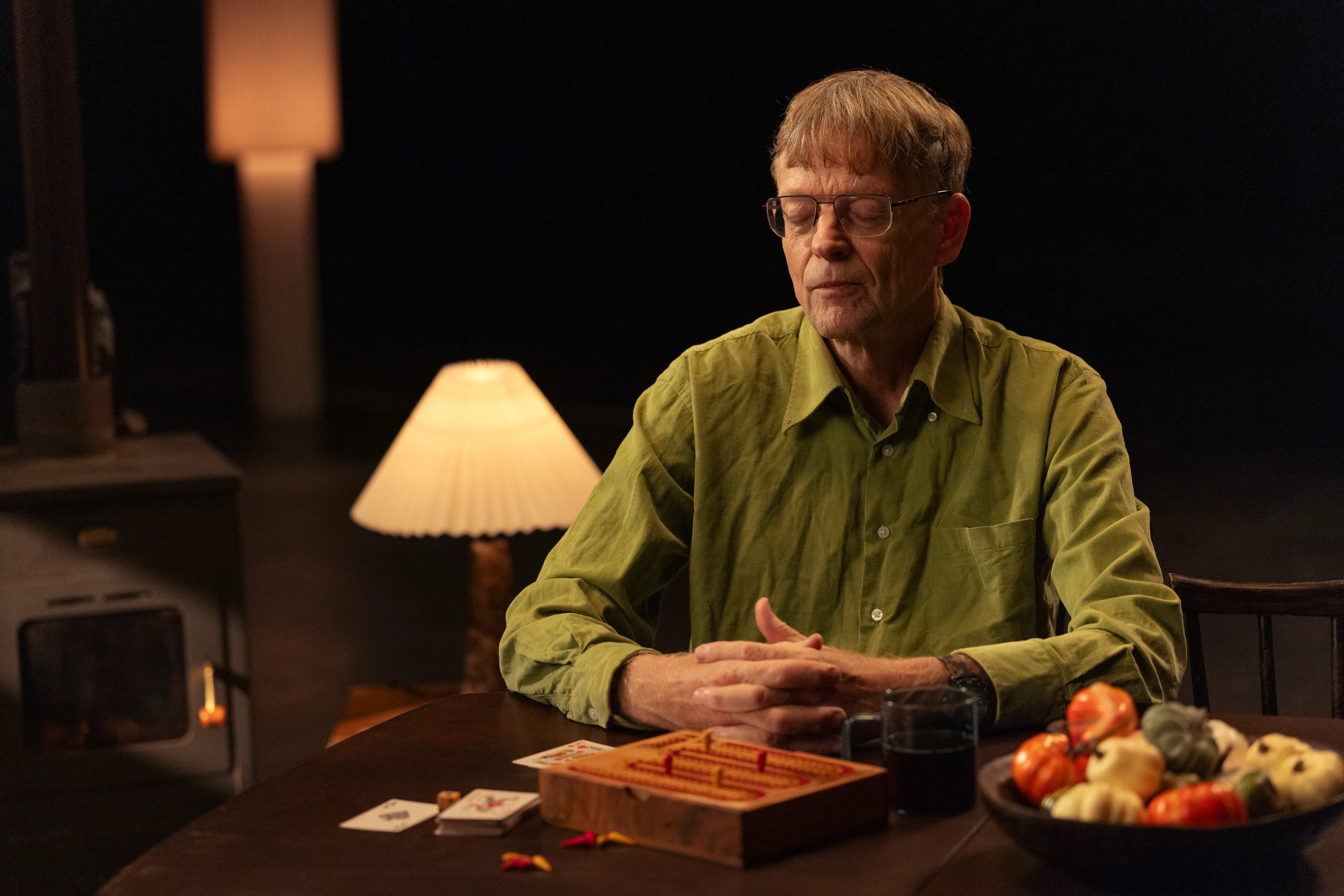 Op de afbeelding zie je een man aan tafel. Op tafel staan een fruitschaal en een spelletje. Op de achtergrond staan een lamp en een houtkachel.