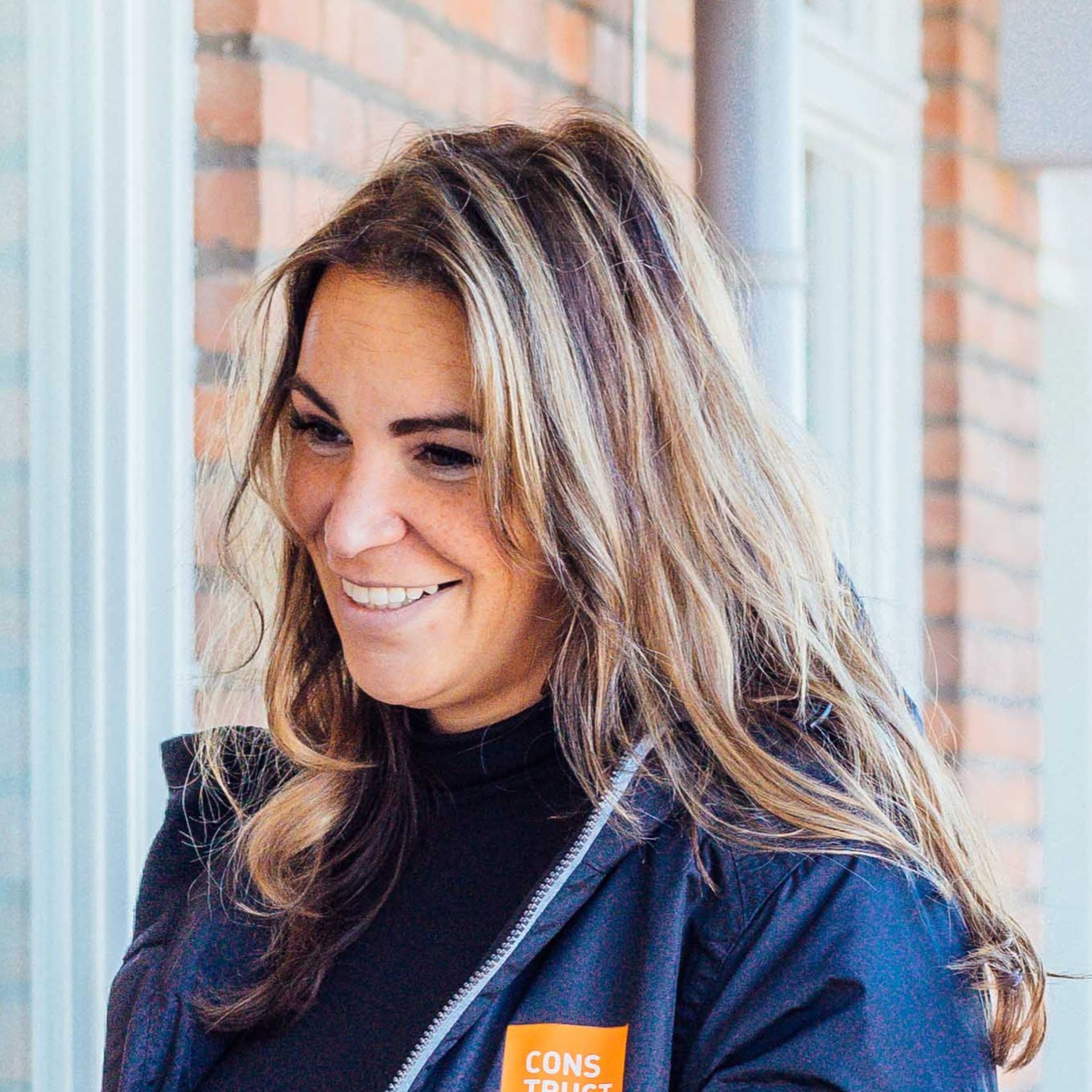 Portretfoto van een vrouw met bruin-blond haar tot op haar schouders. Ze lacht en draagt een blauw jack, met een oranje, rechthoekig logo van het bedrijf Constructif